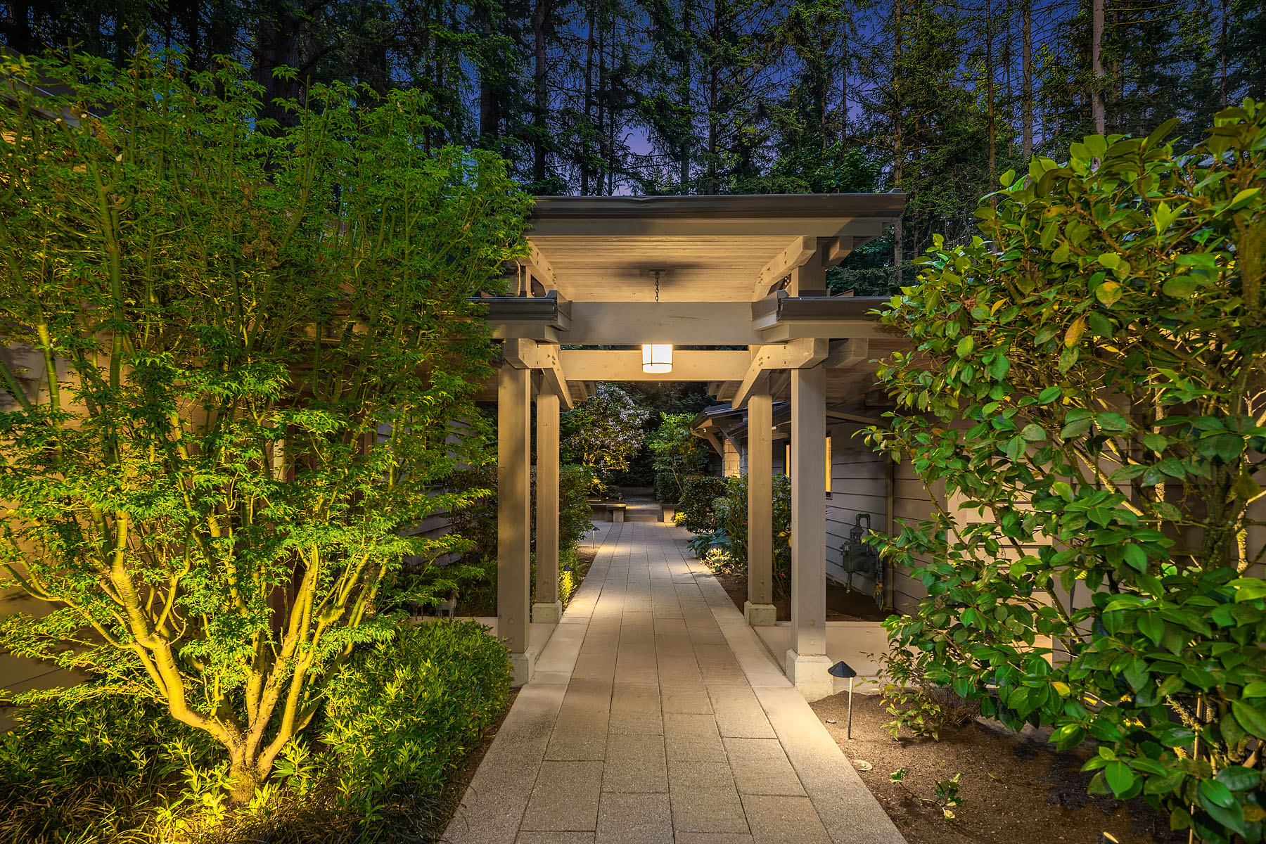 A wood-and-stone entry pergola lit at twilight, framed by maples