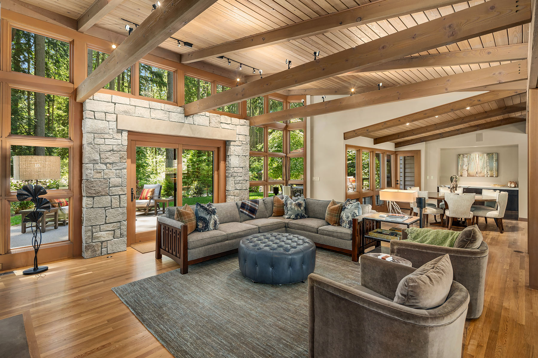 Great room with stone column, vaulted cedar ceiling, exposed beams, and forest beyond the windows