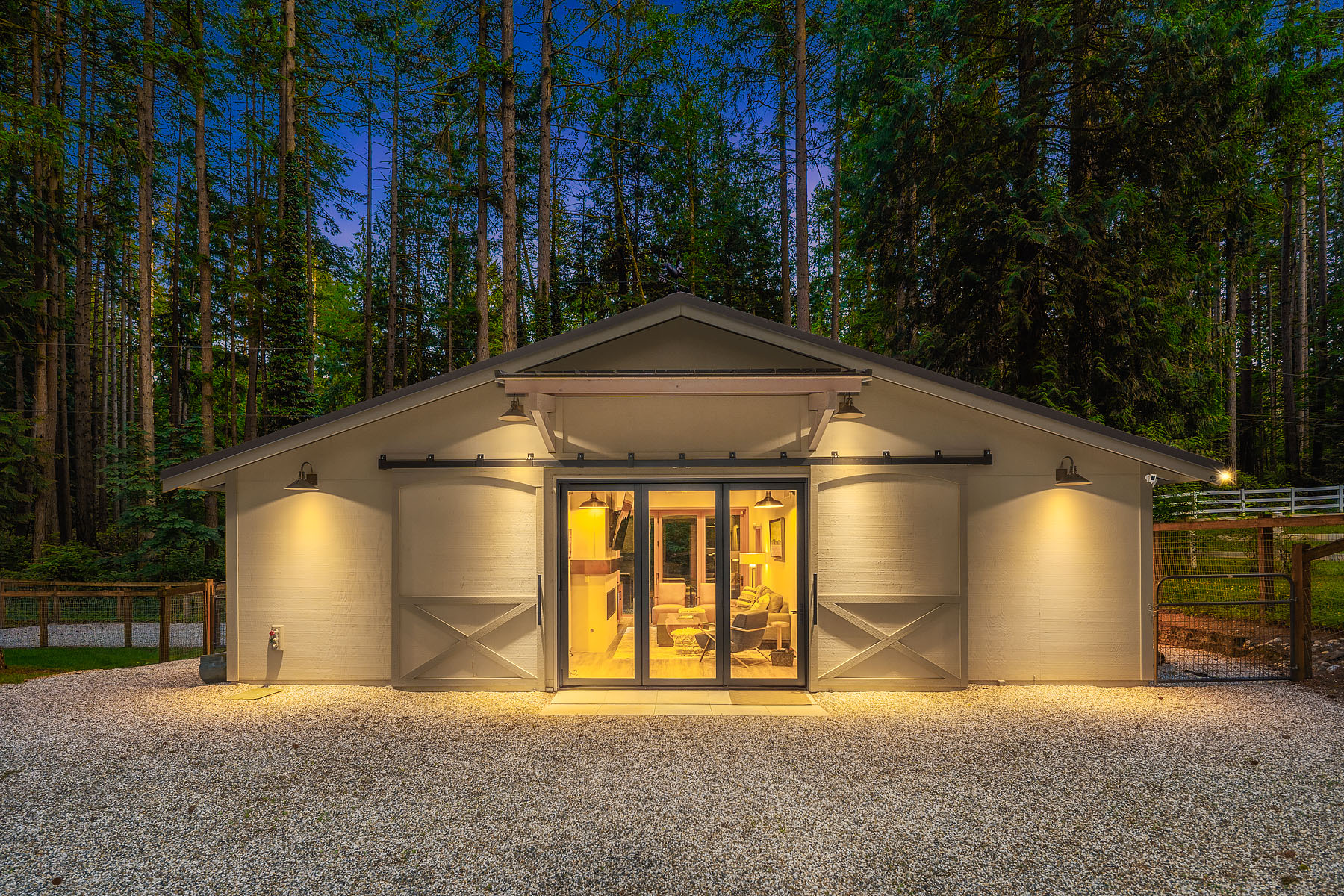 The detached barn at twilight, front-on, lit through center doors with deep cedars rising behind