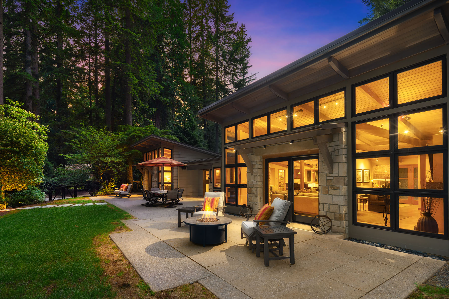 Twilight on the entertainment patio with a fire feature, stone column, and the home glowing through the cedars